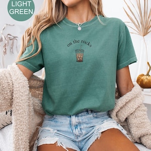 Puede incluir: Camiseta verde claro con el texto "on the rocks" y una ilustraci&oacute;n de caf&eacute; helado. La camiseta de manga corta tiene cuello redondo. La modelo lleva un pantal&oacute;n corto vaquero y una rebeca color crema.