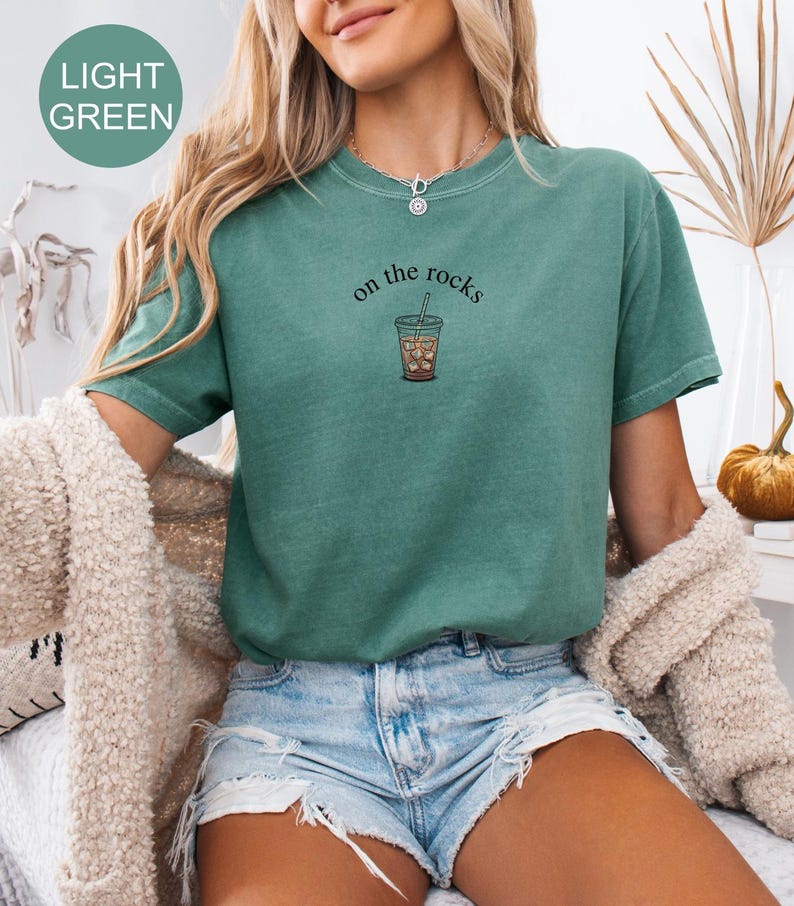Puede incluir: Camiseta verde claro con el texto "on the rocks" y una ilustraci&oacute;n de caf&eacute; helado. La camiseta de manga corta tiene cuello redondo. La modelo lleva un pantal&oacute;n corto vaquero y una rebeca color crema.