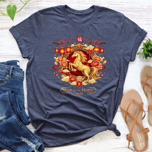 May include: Navy blue t-shirt featuring a golden horse design with red and gold accents, lanterns, and floral elements. The text "Year of the Horse 2026" is displayed below the horse illustration. The shirt is a crew neck.