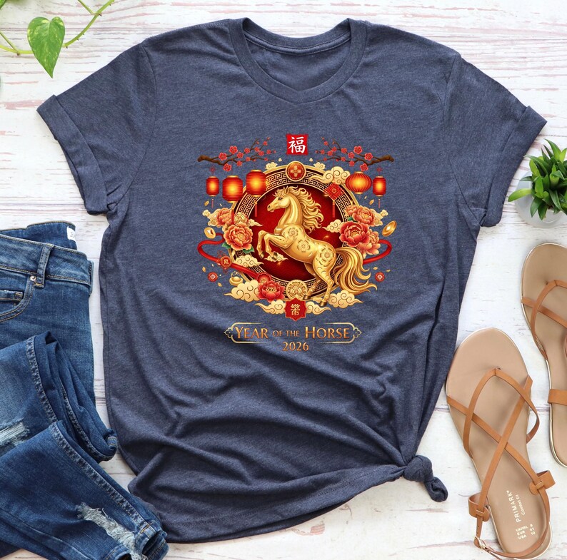 May include: Navy blue t-shirt featuring a golden horse design with red and gold accents, lanterns, and floral elements. The text "Year of the Horse 2026" is displayed below the horse illustration. The shirt is a crew neck.