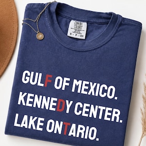 May include: A navy blue short-sleeve t-shirt with white text that reads GULF OF MEXICO, KENNEDY CENTER, and LAKE ONTARIO. The letters F, D, and T are highlighted in red. The shirt features a Comfort Colors brand label on the inner collar.