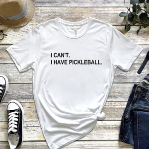 May include: White t-shirt with black text that reads "I CAN'T. I HAVE PICKLEBALL."