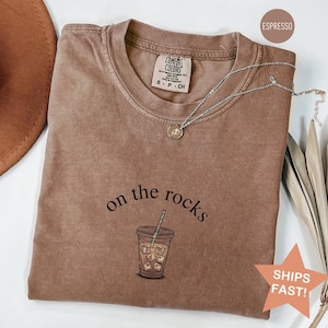 Puede incluir: Camiseta color espresso con la frase "on the rocks" sobre un gr&aacute;fico de caf&eacute; helado. La camiseta tiene cuello redondo y mangas cortas. Un collar plateado con un colgante est&aacute; sobre la camiseta.