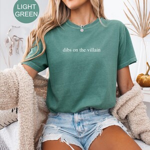 Op de afbeelding: Lichtgroen T-shirt met de witte tekst "dibs on the villain". Het shirt heeft korte mouwen en een relaxte pasvorm. Het model draagt een spijkerbroekshort en een cr&egrave;mekleurig vest.