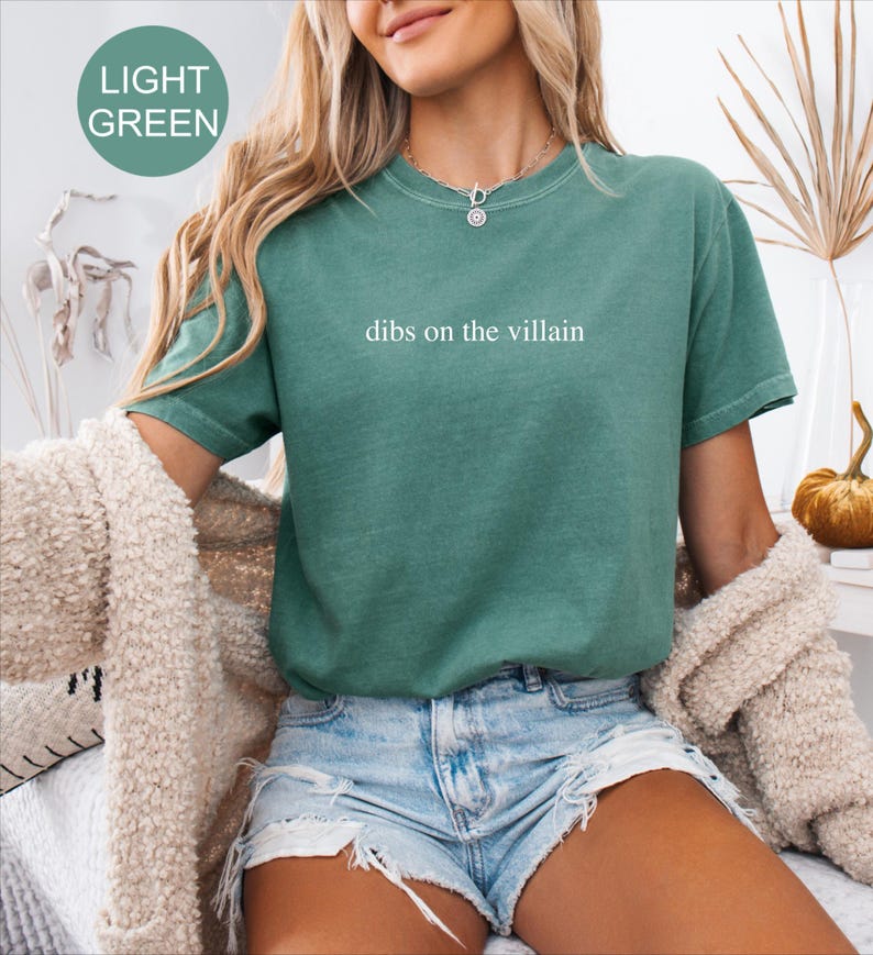 Op de afbeelding: Lichtgroen T-shirt met de witte tekst "dibs on the villain". Het shirt heeft korte mouwen en een relaxte pasvorm. Het model draagt een spijkerbroekshort en een cr&egrave;mekleurig vest.
