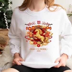 May include: White sweatshirt featuring a golden horse design within a circular frame. The design includes red and gold accents, lanterns, and the text "Year of the Horse 2020". The sweatshirt is a crew neck style.