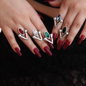 Op de afbeelding: Zilverkleurige ringen met gekleurde edelsteenaccenten. De ringen hebben een V-vormig ontwerp met een centrale ovale steen in rood, helder, groen, blauw en zwart. De ringen worden getoond op een hand met lange, bordeauxrode nagels.