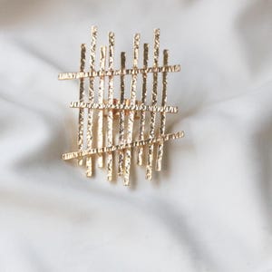 May include: A gold-toned metal ring with a geometric design. The ring is made of a series of thin, textured bars that are arranged in a grid pattern.