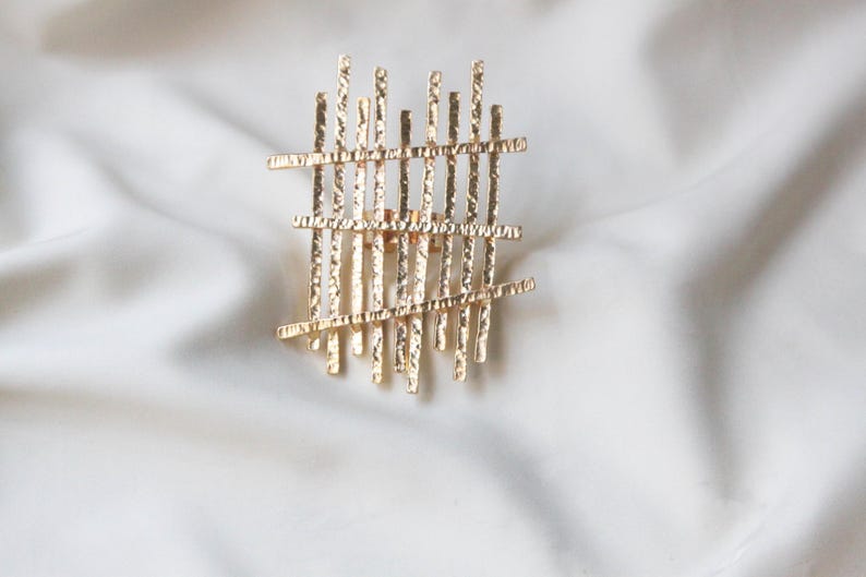 May include: A gold-toned metal ring with a geometric design. The ring is made of a series of thin, textured bars that are arranged in a grid pattern.