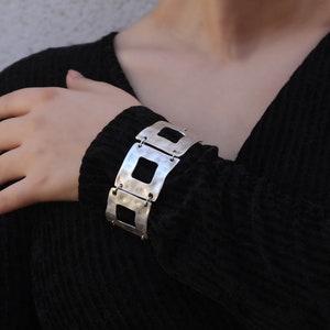 May include: A silver bracelet with a geometric design. The bracelet is made of multiple connected squares with cutouts.