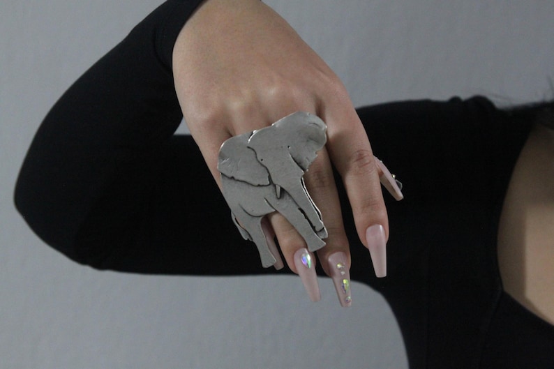 Puede incluir: Un anillo de plata con un dise&ntilde;o de elefante detallado. El anillo se lleva en el dedo &iacute;ndice de una mano con u&ntilde;as largas de color rosa claro.