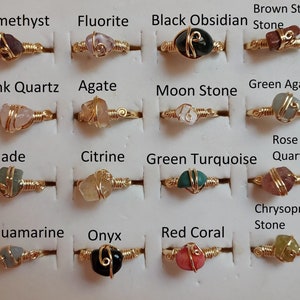 Puede incluir: Una colección de 16 anillos envueltos en oro con gemas de diferentes colores. Los anillos están dispuestos en cuatro filas de cuatro, con cada fila etiquetada con el nombre de la gema. Las gemas incluyen amatista, fluorita, obsidiana negra, piedra estrella marrón, cuarzo rosa, ágata, piedra lunar, ágata verde, jade, citrino, turquesa verde, cuarzo rosa, aguamarina, ónix, coral rojo y crisoprasa.