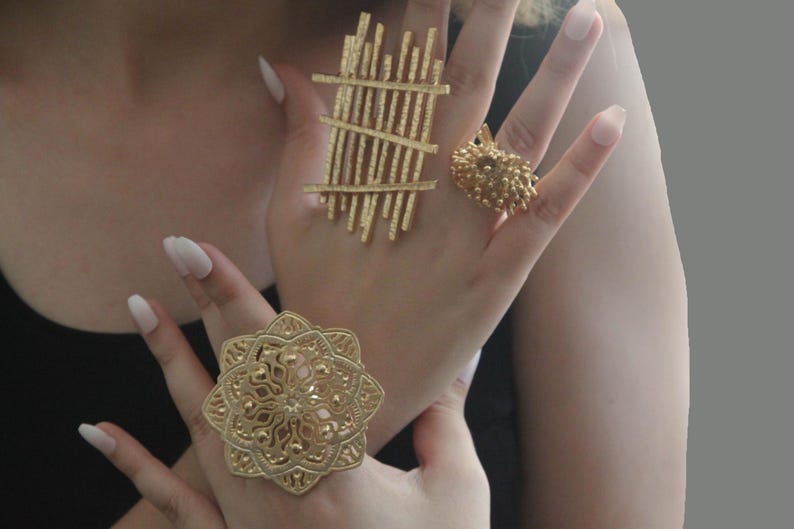 May include: Three gold rings with intricate designs. The first ring has a geometric pattern of vertical and horizontal lines. The second ring has a floral design. The third ring has a cluster of small, round shapes.
