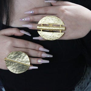 Può includere: Due anelli dorati con una texture martellata e una barra orizzontale al centro. Gli anelli sono indossati sull'indice e sul medio.