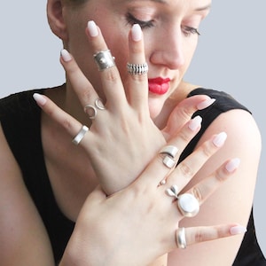 Puede incluir: Una mano de mujer con varios anillos de plata, incluyendo un anillo grande con una piedra blanca. Los anillos están en diferentes dedos y están apilados unos sobre otros.