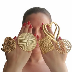 Puede incluir: Una colección de anillos dorados con varios diseños, incluyendo una forma de corazón, un cuadrado texturizado y un diseño circular con círculos concéntricos. Los anillos se muestran en las manos de una persona con esmalte de uñas rojo.