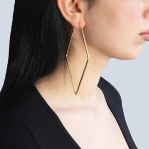 Pode incluir: Brincos dourados em forma de diamante. Os brincos são grandes e pendem do lóbulo da orelha. A modelo está vestindo um top preto. O fundo é azul claro.