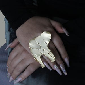 Puede incluir: Un anillo de elefante de color dorado con una superficie texturizada. El anillo se lleva en un dedo con u&ntilde;as largas de color rosa claro.