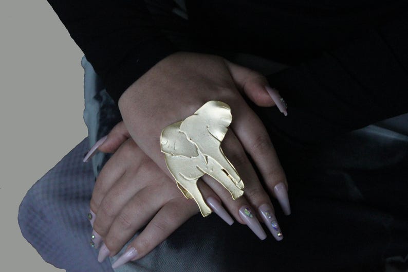 Puede incluir: Un anillo de elefante de color dorado con una superficie texturizada. El anillo se lleva en un dedo con u&ntilde;as largas de color rosa claro.