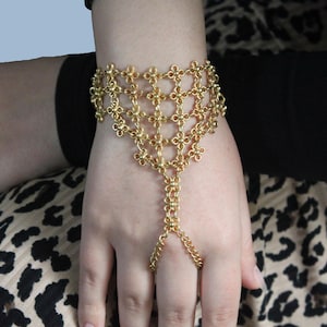 Könnte beinhalten: Ein goldfarbenes Handkettenarmband. Das Armband hat ein florales Design am Handgelenk und eine Kette, die mit einem Ring verbunden ist. Der Schmuck wird an einer Hand mit schwarzem Ärmel und einem Oberteil mit Leopardenmuster im Hintergrund getragen.