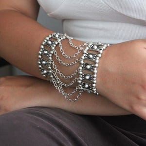 Puede incluir: Una pulsera de tono plateado con dos bandas anchas de eslabones rectangulares conectadas por cinco cadenas plateadas finas y drapeadas. Los eslabones rectangulares están decorados con pequeños tachones circulares en relieve, creando un diseño estructurado.