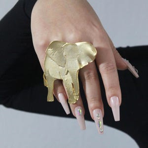 Puede incluir: Un anillo de elefante de color dorado con un dise&ntilde;o detallado. El anillo se lleva en un dedo con u&ntilde;as largas de color rosa claro.