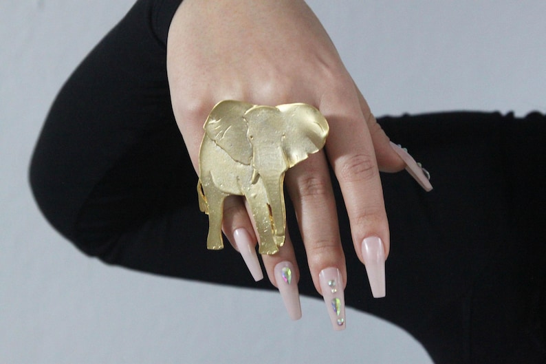 Puede incluir: Un anillo de elefante de color dorado con un dise&ntilde;o detallado. El anillo se lleva en un dedo con u&ntilde;as largas de color rosa claro.