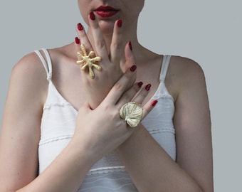 May include: A woman's hands wearing two gold rings. One ring is a large, abstract, starburst design. The other ring is a leaf-shaped design. The woman is wearing a white tank top.