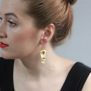 Può includere: Orecchini dorati con un designo a forma di viso. Gli orecchini sono pendenti e hanno una superficie testurizzata.