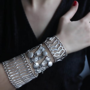 Pode incluir: Um bracelete de prata com múltiplas camadas de designs intrincados, incluindo uma fileira de pequenos círculos, uma camada texturizada e uma camada de formas de coração.