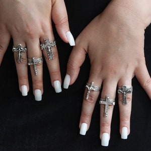 Puede incluir: Las manos de una persona con varios anillos de plata con diseños de cruces. Los anillos se llevan en diferentes dedos y todos son de diferentes tamaños y estilos.