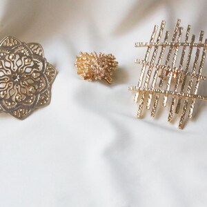May include: Three gold-toned metal hair clips with different designs. One clip is a circular design with intricate details, another is a cluster of small, textured pieces, and the third is a geometric design with straight lines.