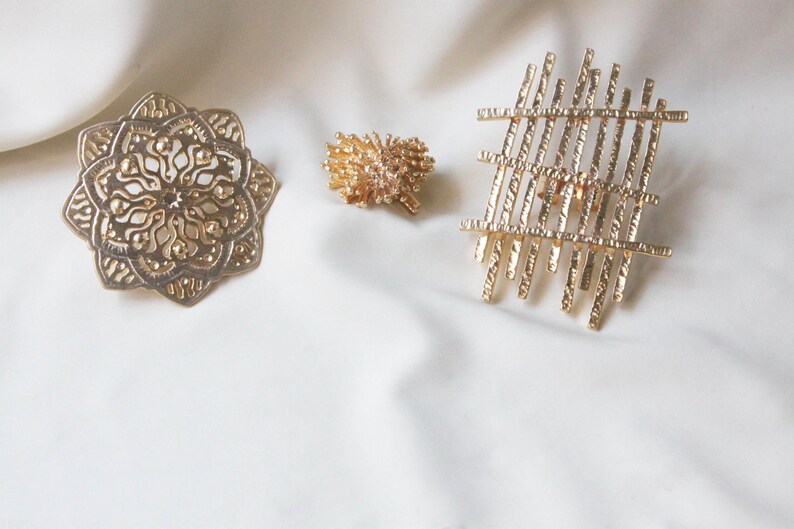 May include: Three gold-toned metal hair clips with different designs. One clip is a circular design with intricate details, another is a cluster of small, textured pieces, and the third is a geometric design with straight lines.