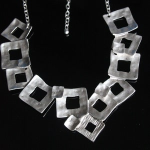 May include: A silver necklace with a chain and a series of interconnected square-shaped pendants. The pendants are open and have a hammered texture.