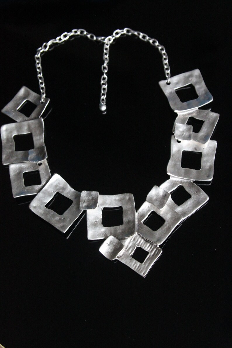 May include: A silver necklace with a chain and a series of interconnected square-shaped pendants. The pendants are open and have a hammered texture.