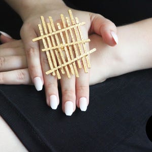 May include: A gold-toned ring with a geometric design. The ring is made of thin, interwoven metal bars that create a grid-like pattern.