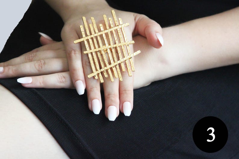 May include: A gold-toned ring with a geometric design. The ring is made of thin, interwoven metal bars that create a grid-like pattern.