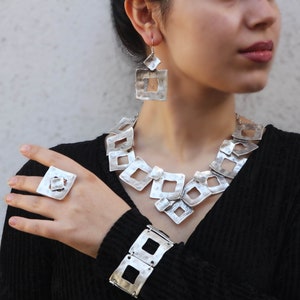 May include: A silver jewelry set with a chunky necklace, bracelet, ring, and earrings. The jewelry features a geometric design with square shapes.