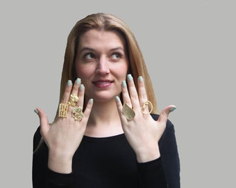 May include: A woman's hands wearing several gold rings. The rings are all different styles, including a leaf design, a geometric design, and a ring with a small charm. The woman's nails are painted a light green color.