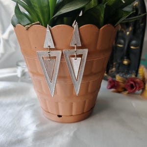 May include: A pair of silver geometric triangle earrings with a layered design. The earrings are hanging from a terracotta pot.