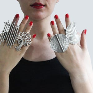 May include: A set of three silver rings featuring a tree design, a circular design, and a spiral design. The rings are worn on the fingers of a person's hand.