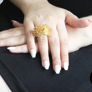 May include: A gold ring featuring a hedgehog design. The ring is worn on a finger with white nail polish.