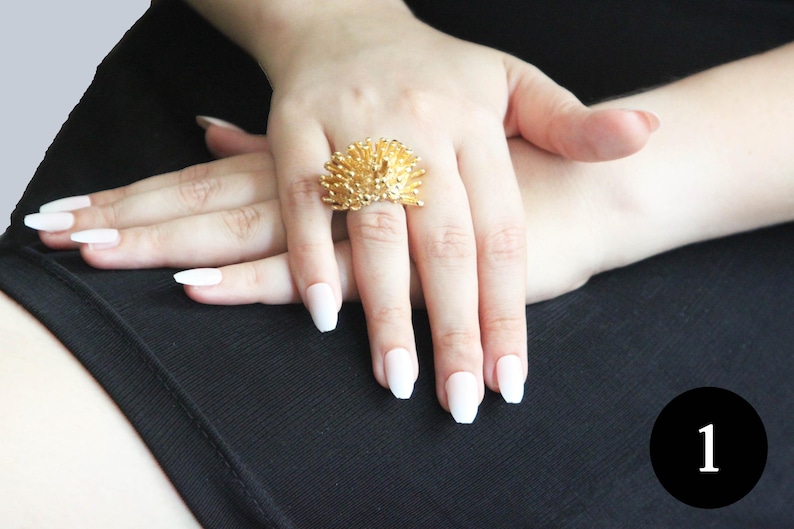May include: A gold ring featuring a hedgehog design. The ring is worn on a finger with white nail polish.