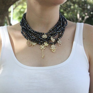 May include: A layered black choker necklace with gold charms, including a key, a clover, a hand, a butterfly, and a circle with a dragonfly.