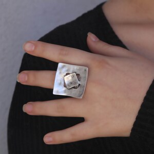 May include: A silver ring with a square design featuring a smaller square cutout in the center. The ring has a textured, hammered finish.