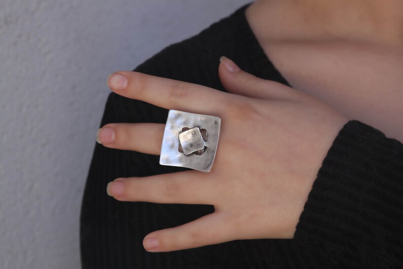 May include: A silver ring with a square design featuring a smaller square cutout in the center. The ring has a textured, hammered finish.
