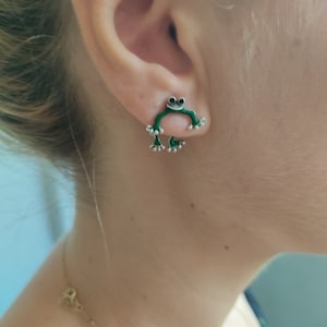 May include: Green frog shaped earrings with silver accents. The frog is sitting on its hind legs with its front legs outstretched.