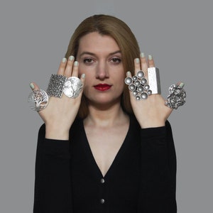 May include: A woman wearing five silver rings on her fingers. The rings are all different designs, including a tree, a geometric pattern, a cluster of circles, and a textured design.