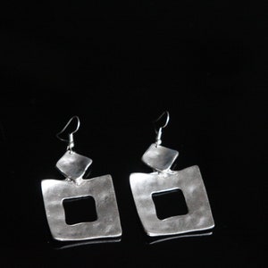 May include: A pair of silver dangle earrings with a square shape and a smaller square cutout in the center. The earrings have a hammered texture and a slightly irregular shape.