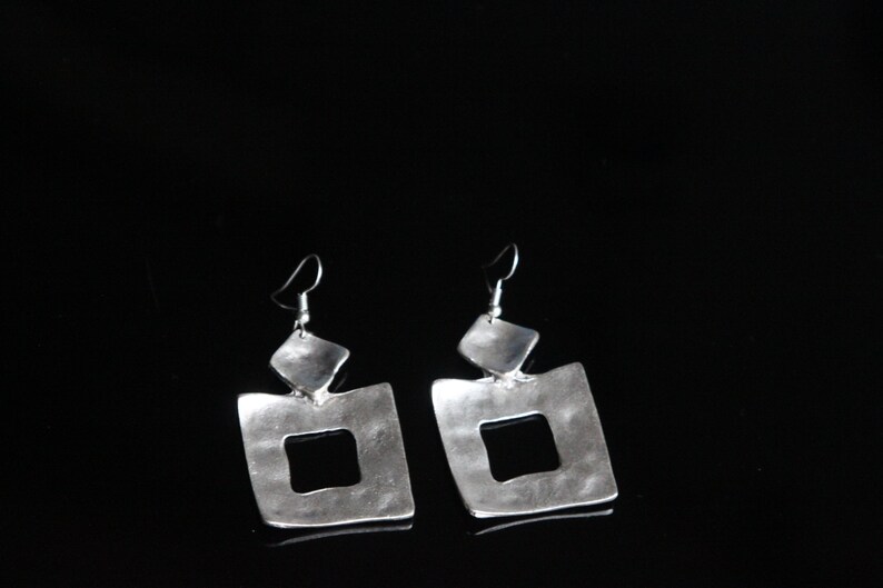 May include: A pair of silver dangle earrings with a square shape and a smaller square cutout in the center. The earrings have a hammered texture and a slightly irregular shape.